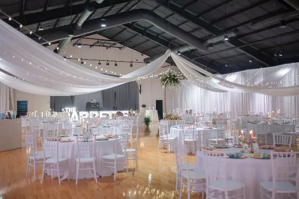 Interior of a large wedding venue with white draped ceilings, string lights, and round tables set with white linens and chairs.