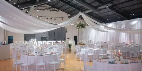 Interior of a large wedding venue with white draped ceilings, string lights, and round tables set with white linens and chairs.