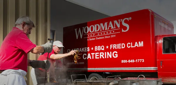 Two chefs in red shirts preparing lobster on a grill in front of a red Woodman's of Essex catering truck.