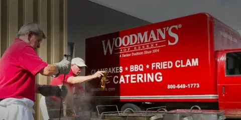 Two chefs in red shirts preparing lobster on a grill in front of a red Woodman's of Essex catering truck.