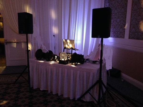 A professional DJ booth setup featuring a laptop, audio mixer, and large speakers on a table in a dimly lit room.