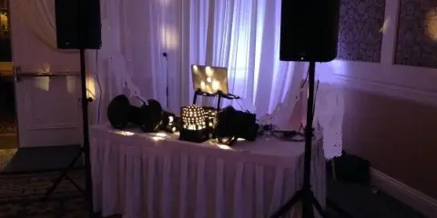 A professional DJ booth setup featuring a laptop, audio mixer, and large speakers on a table in a dimly lit room.