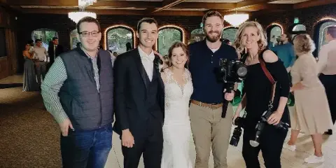 A wedding photography team posing with a bride and groom in a banquet hall.