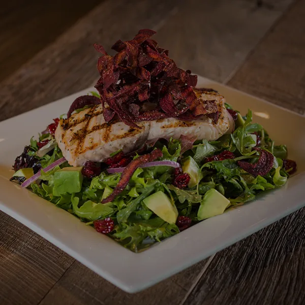 A plated dish featuring grilled fish on a bed of mixed greens with avocado and bacon garnish.