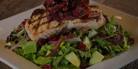 A plated dish featuring grilled fish on a bed of mixed greens with avocado and bacon garnish.