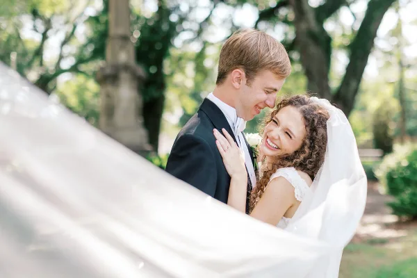 A smiling bride and groom embracing outdoors with a blurred white veil in the foreground.