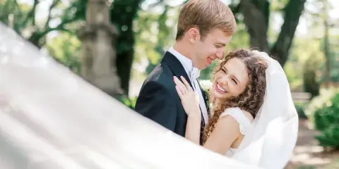 A smiling bride and groom embracing outdoors with a blurred white veil in the foreground.