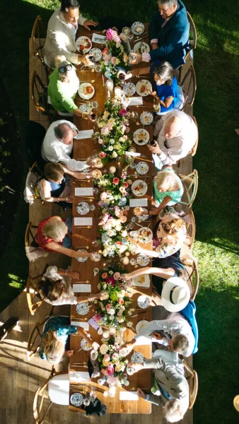 An overhead view of a long wooden dining table set with a floral centerpiece and surrounded by guests at an outdoor event.