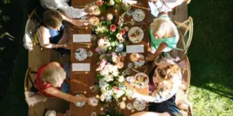 An overhead view of a long wooden dining table set with a floral centerpiece and surrounded by guests at an outdoor event.