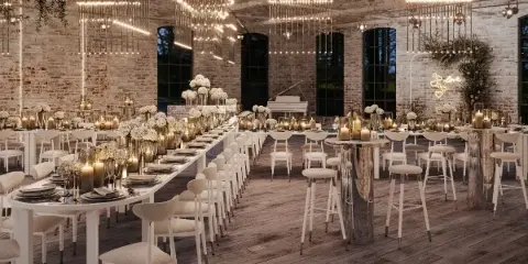 Styled event venue interior with long white banquet tables, upholstered chairs, bar stools, candles, floral centerpieces, and hanging light installations.