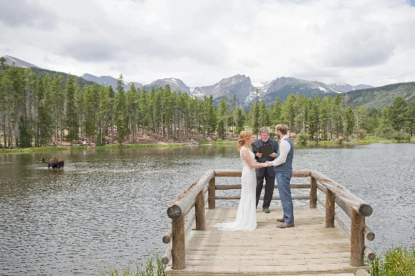 A wedding officiant performing a ceremony for a bride and groom on a wooden dock overlooking a lake with mountains in the background.