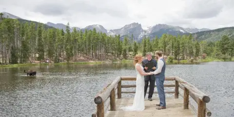 A wedding officiant performing a ceremony for a bride and groom on a wooden dock overlooking a lake with mountains in the background.