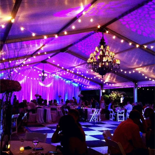 A wedding reception tent interior illuminated with purple event lighting, chandeliers, and string lights.