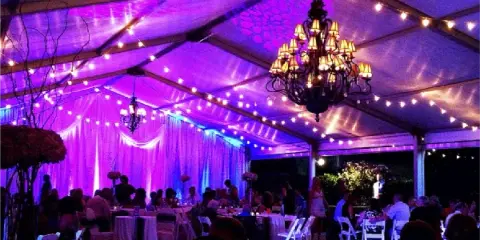 A wedding reception tent interior illuminated with purple event lighting, chandeliers, and string lights.