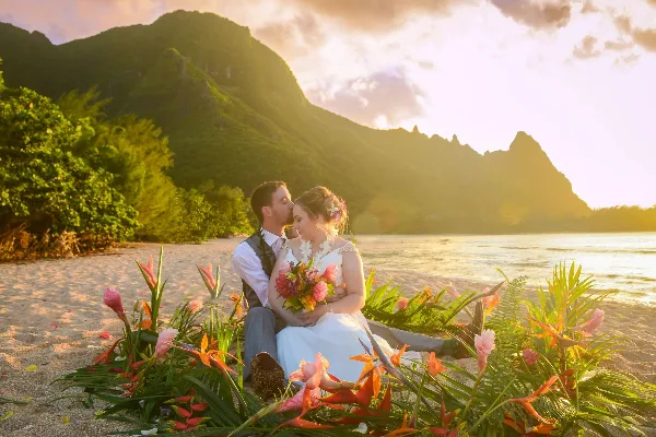 A bride and groom sitting on a sandy beach surrounded by tropical flowers with a mountain backdrop at sunset.