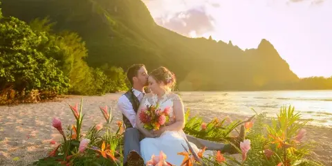 A bride and groom sitting on a sandy beach surrounded by tropical flowers with a mountain backdrop at sunset.