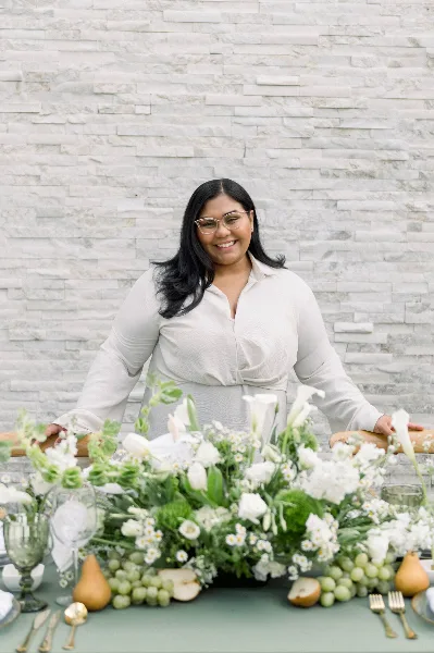 A smiling woman in a white blouse stands behind a wedding table decorated with white flowers, greenery, and fruit.