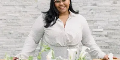 A smiling woman in a white blouse stands behind a wedding table decorated with white flowers, greenery, and fruit.