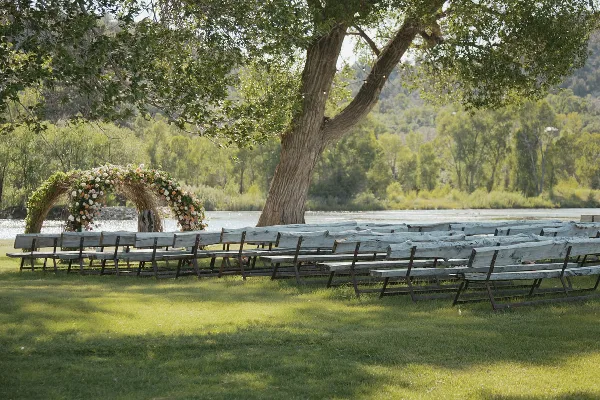 Outdoor wedding ceremony setup with a floral willow arbor and wooden benches on a grassy lawn beside a lake.
