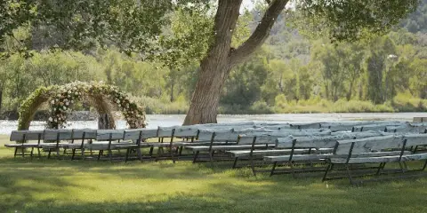 Outdoor wedding ceremony setup with a floral willow arbor and wooden benches on a grassy lawn beside a lake.