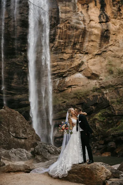 A bride and groom kissing in front of a large waterfall at a rocky outdoor venue.