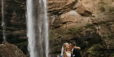 A bride and groom kissing in front of a large waterfall at a rocky outdoor venue.