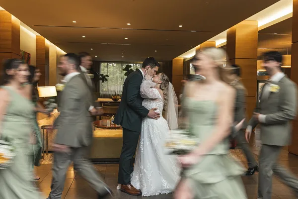A bride and groom kiss in the center of a hotel lobby while their wedding party walks around them, captured with a motion blur effect.