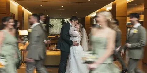 A bride and groom kiss in the center of a hotel lobby while their wedding party walks around them, captured with a motion blur effect.