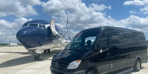 A black Mercedes-Benz Sprinter van parked on an airport tarmac next to a private jet.