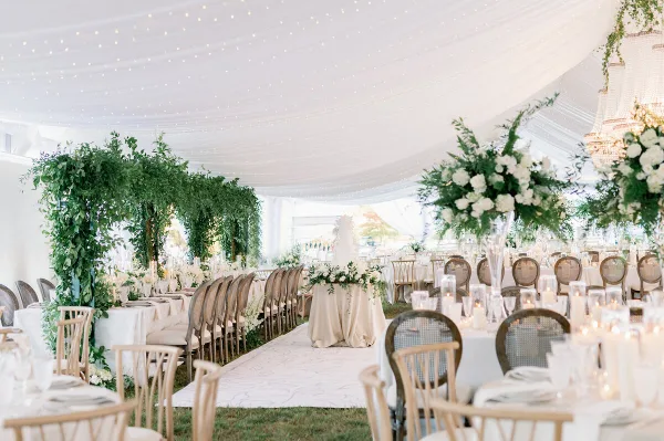 A luxury wedding reception setup inside a white tent featuring a floral arch, long banquet tables with white linens, and a ceiling draped with string lights.
