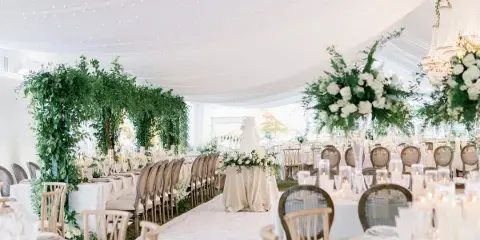 A luxury wedding reception setup inside a white tent featuring a floral arch, long banquet tables with white linens, and a ceiling draped with string lights.