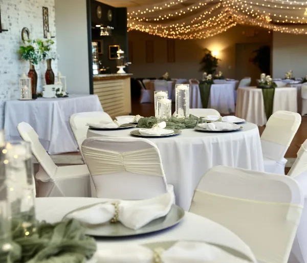 Interior view of a wedding reception hall with round tables, white linens, and string lights.