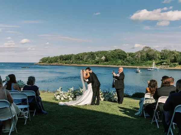 A bride and groom share a kiss during a wedding ceremony on a grassy lawn overlooking a harbor with boats and a wooded coastline in the background.