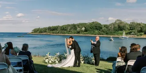 A bride and groom share a kiss during a wedding ceremony on a grassy lawn overlooking a harbor with boats and a wooded coastline in the background.