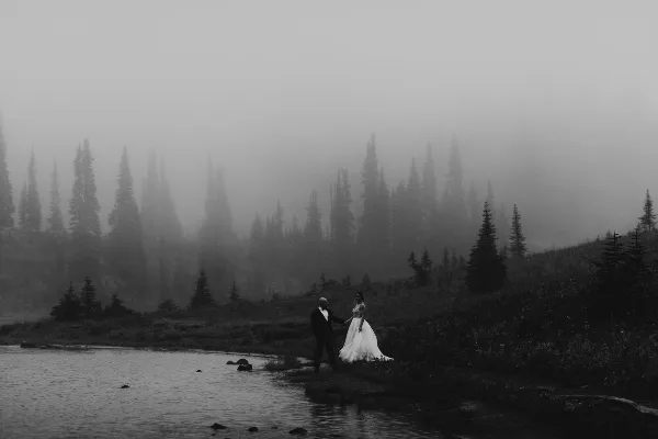 A black and white photograph of a bride and groom standing by a lake in a foggy, mountainous forest setting.