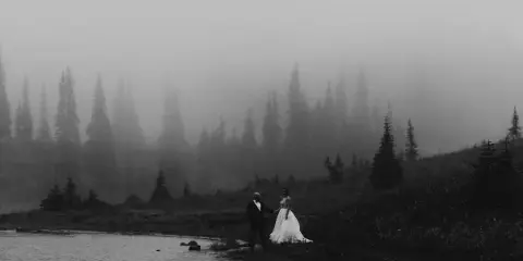 A black and white photograph of a bride and groom standing by a lake in a foggy, mountainous forest setting.