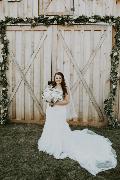 A bride in a white wedding dress holding a bouquet stands in front of a rustic wooden barn decorated with floral garlands.