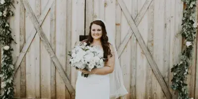 A bride in a white wedding dress holding a bouquet stands in front of a rustic wooden barn decorated with floral garlands.