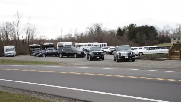 A fleet of black SUVs, limousines, and white buses parked in a gravel lot beside a road.