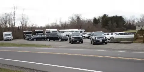 A fleet of black SUVs, limousines, and white buses parked in a gravel lot beside a road.