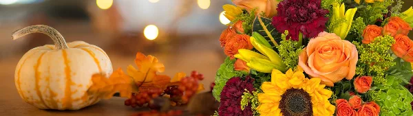 Fresh fall floral arrangement featuring orange roses, sunflowers, and lilies beside a small pumpkin and autumn leaves.