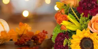 Fresh fall floral arrangement featuring orange roses, sunflowers, and lilies beside a small pumpkin and autumn leaves.