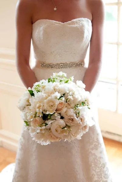 A close-up of a bride holding a large, round bouquet of white and cream roses and peonies.