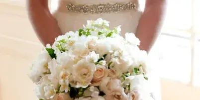 A close-up of a bride holding a large, round bouquet of white and cream roses and peonies.
