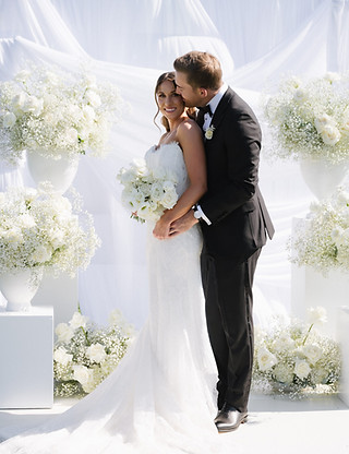 A bride and groom embracing at a wedding ceremony with white floral arrangements.