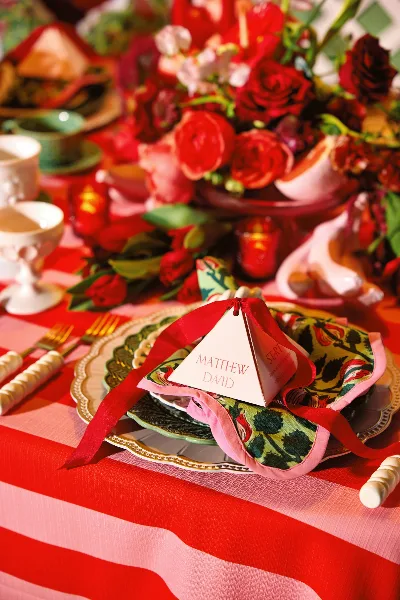 A close-up of a styled wedding table setting featuring stacked vintage plates, floral napkins, and gold flatware on a red and pink striped tablecloth.