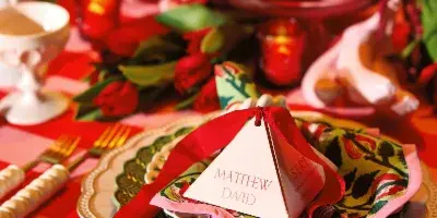 A close-up of a styled wedding table setting featuring stacked vintage plates, floral napkins, and gold flatware on a red and pink striped tablecloth.