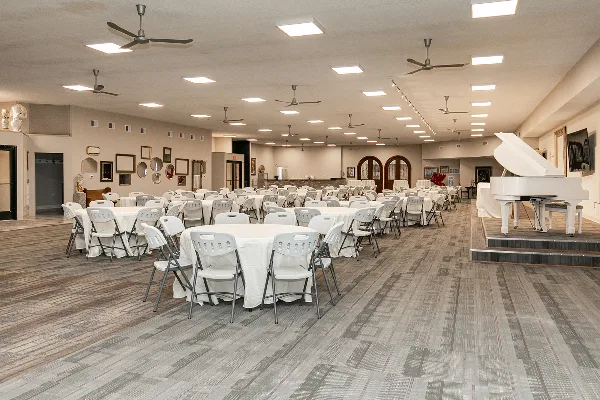 Interior view of a large event hall featuring round tables with white linens, white folding chairs, and a white grand piano on a raised stage.