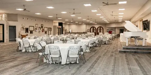 Interior view of a large event hall featuring round tables with white linens, white folding chairs, and a white grand piano on a raised stage.