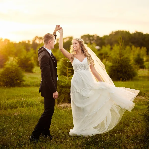 A bride and groom dancing in a grassy field at sunset, with the bride's dress and veil flowing in the wind.
